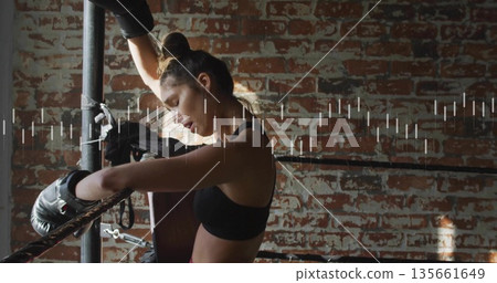 Leaning boxer wearing black sports bra and gloves, resting on ropes at gym, with brick wall 135661649