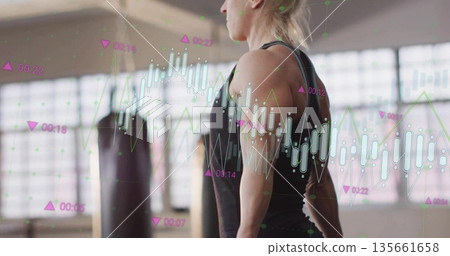 Standing woman in black top, leggings in gym with punching bags, displaying data charts, copy space Standing woman in black top, leggings in gym with punching bags, displaying data charts, copy space 135661658