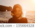 Woman sunset beach portrait featuring a happy traveler wearing a beanie near the sea arch at golden hour. 135664328