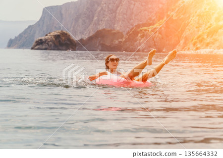 Woman sea inflatable laughing while floating on a pink ring in the ocean near rocky cliffs during sunset 135664332