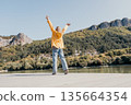 Woman in Yellow Jacket Raising Arms on a Wooden Dock 135664354
