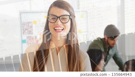 Smiling woman wearing blouse and glasses in office, with desk, sticky-note whiteboard, code overlay 135664943