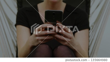 Holding smartphone, woman wearing dark top and leggings typing at home in hanging chair with ropes 135664980