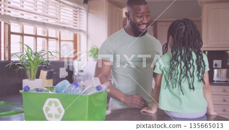 Smiling father teaching daughter sorting plastic bottles into green recycling bin in kitchen 135665013