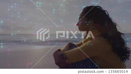 Sitting adult woman hugging knees on sandy beach at sunset, with yellow sweater, copy space 135665014