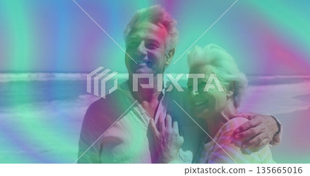 Smiling senior couple posing on sandy shoreline wearing light clothing, with waves rolling behind Smiling senior couple posing on sandy shoreline wearing light clothing, with waves rolling behind 135665016