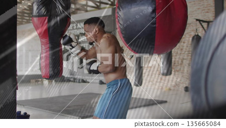 Striking shirtless boxer wearing blue shorts and gloves at boxing gym, with heavy bag, water bottle 135665084