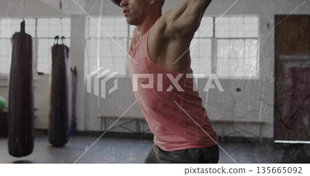 Performing overhead press muscular man inside gym, with hanging punching bags and exercise ball 135665092