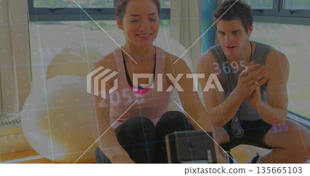 Woman using rower in gym with stopwatch, digital metric overlay and kneeling trainer guiding her 135665103