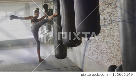 Athlete wearing sports bra and boxing gloves delivering kick in gym with punching bags, copy space 135665149
