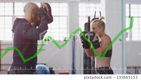 Punching female boxer wearing gloves with coach holding focus mitts in training studio, with graph 135665153