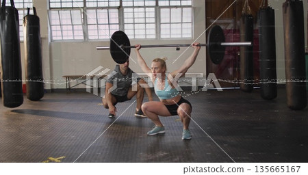 Performing overhead squat with barbell, woman in sportswear at gym with trainer spotting 135665167