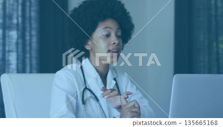 Speaking female doctor gesturing toward laptop in consultation room, with stethoscope on desk Speaking female doctor gesturing toward laptop in consultation room, with stethoscope on desk 135665168