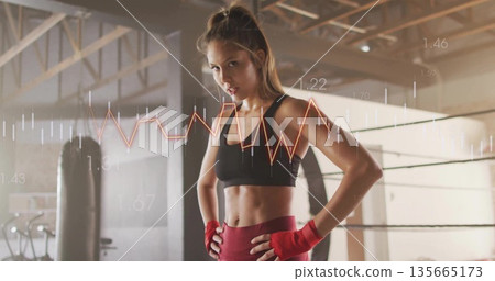 Standing female athlete flexing in gym punching bag wearing red hand wraps showing metrics overlay 135665173