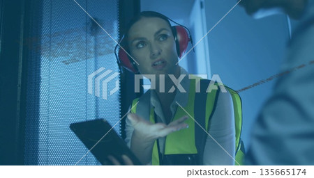 Gesturing mid adult female worker holding tablet in equipment room, with safety vest, copy space 135665174