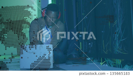 Typing technician wearing red headphones on silver laptop at data center with server racks, cables Typing technician wearing red headphones on silver laptop at data center with server racks, cables 135665183