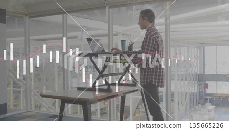 Standing man in plaid shirt using desk converter analyzing charts in open office, with laptop Standing man in plaid shirt using desk converter analyzing charts in open office, with laptop 135665226