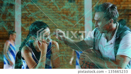 Leaning coach holding basketball, speaking to teen player on gym sideline bench, with team jerseys Leaning coach holding basketball, speaking to teen player on gym sideline bench, with team jerseys 135665233