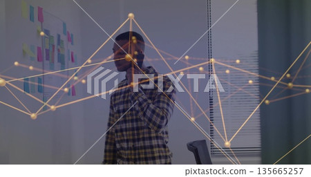 Standing African American man speaking into smartphone in office with sticky notes and data overlay Standing African American man speaking into smartphone in office with sticky notes and data overlay 135665257