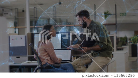 Coworkers discussing data in open-plan office, with dual monitors, laptop and globe hologram Coworkers discussing data in open-plan office, with dual monitors, laptop and globe hologram 135665286