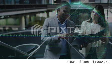 Reviewing suited colleagues checking tablet and smartphone in corporate atrium, with data overlay 135665289