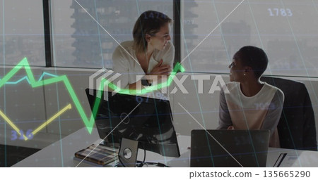 Discussing data, two business-casual women at desk in office, with monitor and financial chart 135665290