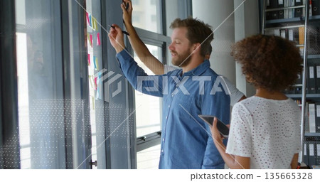 Three colleagues collaborating at glass wall in modern office, using sticky notes, reviewing tablet 135665328