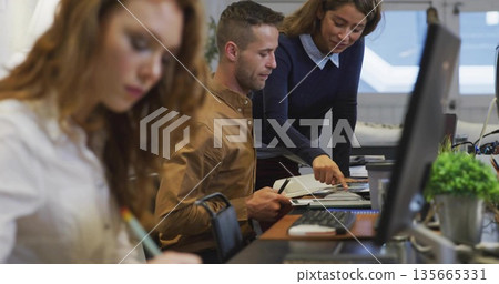 Discussing two colleagues reviewing documents in modern office, with monitors, notebooks and pens 135665331