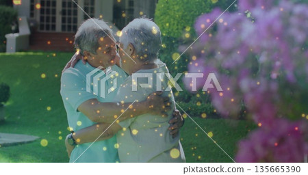 Embracing senior couple touching heads in front yard with smartwatch pink shrub and light particles Embracing senior couple touching heads in front yard with smartwatch pink shrub and light particles 135665390