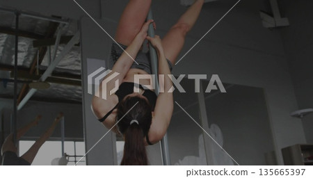 Woman performing inverted pole spin in bra and shorts at fitness studio, with mirrored reflection Woman performing inverted pole spin in bra and shorts at fitness studio, with mirrored reflection 135665397