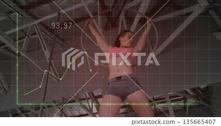 Athlete wearing sports bra, shorts hanging on rings on rig in gym, with performance graph overlay Athlete wearing sports bra, shorts hanging on rings on rig in gym, with performance graph overlay 135665407