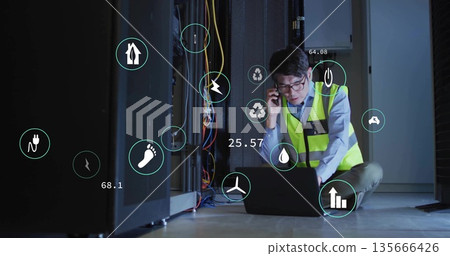 Crouching Korean technician wearing safety vest using phone, laptop in server room with eco icons 135666426