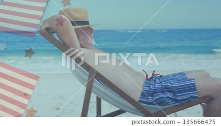 Lounging older man reclining on beach chair at shoreline, with striped swim trunks and straw fedora 135666475