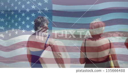 Holding surfboards, senior women in swimsuits watching waves on beach, with American flag overlay 135666488