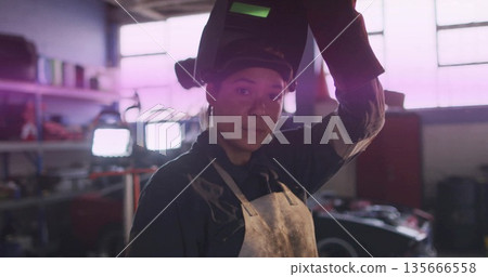 Lifting welding helmet, female welder in workshop wearing leather apron amid LED lamps and shelving 135666558