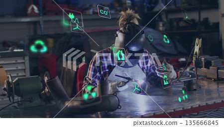 Leaning welder wearing helmet, gloves holding welding rod at workshop, showing sustainability icons 135666845