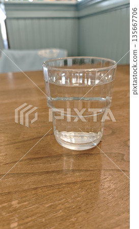 Glass of fresh water standing on a wooden table in a cafe interior with soft natural light. The image represents hydration, simplicity, minimalism, and healthy everyday lifestyle Glass of fresh water standing on a wooden table in a cafe interior with soft natural light. The image represents hydration, simplicity, minimalism, and healthy everyday lifestyle 135667706