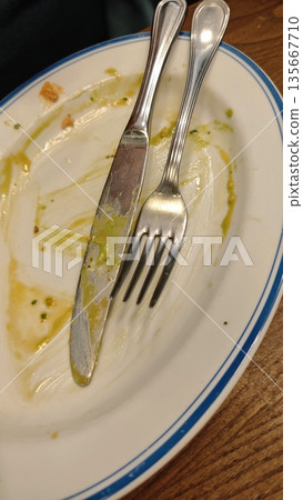 Empty plate with knife and fork showing traces of sauce after a finished meal on a wooden table. The image represents completed dining, satisfaction, minimalism, and the end of a food experience Empty plate with knife and fork showing traces of sauce after a finished meal on a wooden table. The image represents completed dining, satisfaction, minimalism, and the end of a food experience 135667710