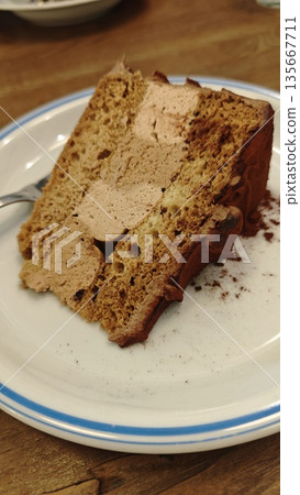 Slice of layered sponge cake with creamy filling served on a white plate on a wooden table in a cafe. The image represents dessert indulgence, sweet treat, coffee break, and cozy cafe lifestyle Slice of layered sponge cake with creamy filling served on a white plate on a wooden table in a cafe. The image represents dessert indulgence, sweet treat, coffee break, and cozy cafe lifestyle 135667711