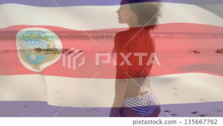 Facing ocean water, woman wearing striped bikini at beach, with tricolor flag overlay, copy space 135667762