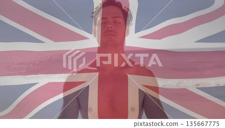 Standing shirtless male wearing trunks closing eyes at beach shoreline, calm sea, UK flag overlay 135667775
