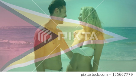 Smiling couple looking at each other wearing white swimwear on beach, with waves and flag overlay Smiling couple looking at each other wearing white swimwear on beach, with waves and flag overlay 135667779