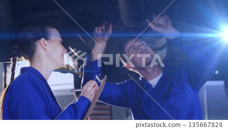 Two mechanics in coveralls inspecting undercarriage on vehicle lift at workshop, with clipboard pen Two mechanics in coveralls inspecting undercarriage on vehicle lift at workshop, with clipboard pen 135667828