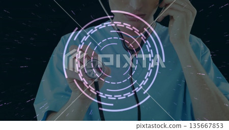 Extending stethoscope, doctor in light blue scrubs before dark backdrop with digital interface 135667853