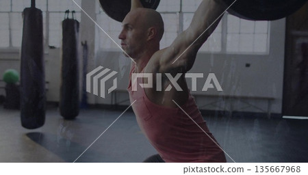 Hoisting Olympic barbell with plates overhead man wearing sportswear in gym with punching bags 135667968