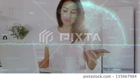 Remote worker typing and holding payment card in kitchen, with laptop, mug, vase and data overlay 135667984