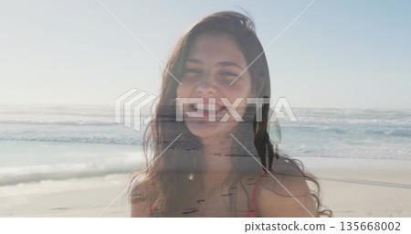 Smiling woman wearing red bikini top at sandy shoreline, with wetsuit surfer carrying surfboard 135668002