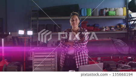 Standing technician in red plaid shirt crossing arms in auto workshop, with laptop and car hood 135668194