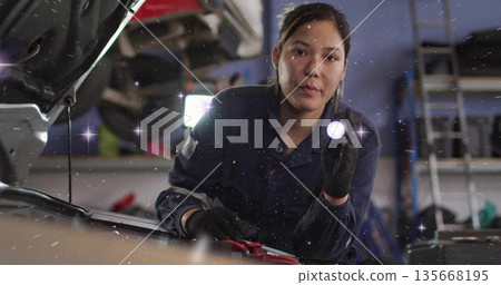 Kneeling woman mechanic in coveralls checking car engine in shop, flashlight lighting jumper clamps Kneeling woman mechanic in coveralls checking car engine in shop, flashlight lighting jumper clamps 135668195