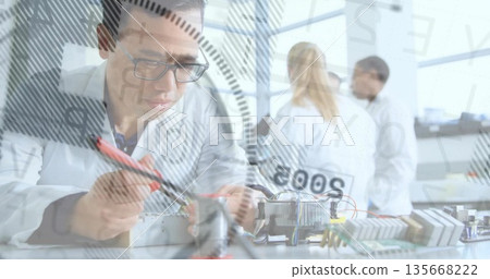 Male researcher in lab coat probing electronics on workbench in lab, with circuit boards and wiring Male researcher in lab coat probing electronics on workbench in lab, with circuit boards and wiring 135668222